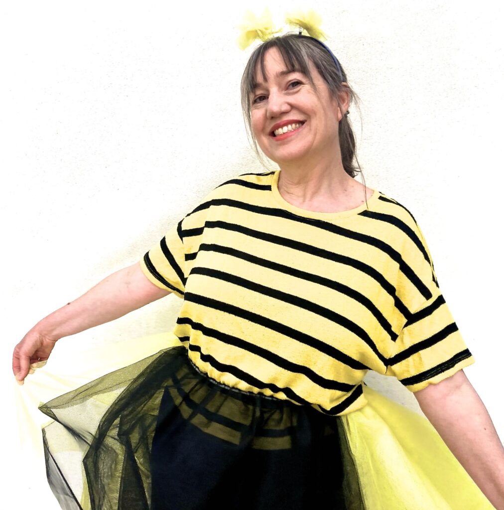 Photo of Alice in a bee costume: yellow and black striped teeshirt with black and yellow tutu and yellow fluffy antennae. She is holding out the yellow sides of the tutu and smiling at the camera.